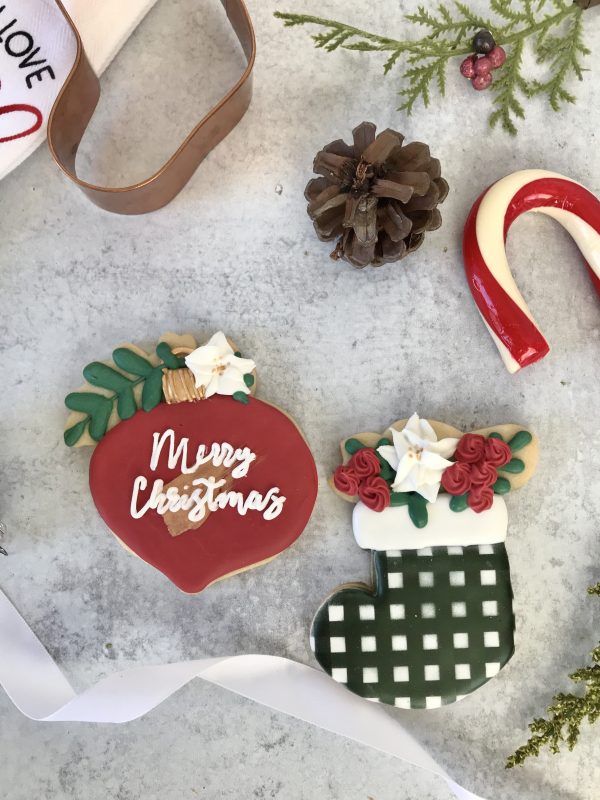 Burgundy ornament with a gold stroke and Merry Christmas written in white with a greenery accent and a poinsettia flower. The stocking is a black and white gingham pattern with royal icing poinsettias in burgundy and white.
