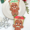 Two decorated gingerbread person sugar cookies displayed vertically on a white surface next to holiday decor, including white jingle bells, a red ornament, and pine greenery. The top cookie, set on a round white plate, features a gingerbread girl with rosy cheeks, a white dress with red heart buttons, and a red hair bow, holding a rectangular banner above her head that reads "Christmas" in green script. The bottom cookie features a gingerbread boy with rosy cheeks, a light green bow tie, and three red buttons, holding a rd banner that reads "Love" in green script.