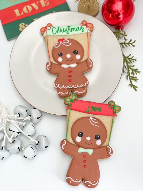 Two decorated gingerbread person sugar cookies displayed vertically on a white surface next to holiday decor, including white jingle bells, a red ornament, and pine greenery. The top cookie, set on a round white plate, features a gingerbread girl with rosy cheeks, a white dress with red heart buttons, and a red hair bow, holding a rectangular banner above her head that reads "Christmas" in green script. The bottom cookie features a gingerbread boy with rosy cheeks, a light green bow tie, and three red buttons, holding a rd banner that reads "Love" in green script.