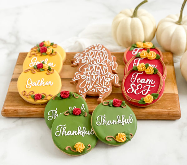 Assorted decorated Thanksgiving sugar cookies featuring autumn-colored icing, festive text like "Gather" and "Thankful," floral piping accents, and a turkey-shaped cookie, set on a wooden platter alongside mini white pumpkins.
