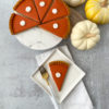 pumpkinpieslice A slice of pumpkin pie on a white square plate next to a gold fork, with the rest of the pie sliced on a marble server alongside small decorative white and yellow pumpkins.