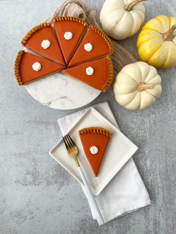 pumpkinpieslice A slice of pumpkin pie on a white square plate next to a gold fork, with the rest of the pie sliced on a marble server alongside small decorative white and yellow pumpkins.