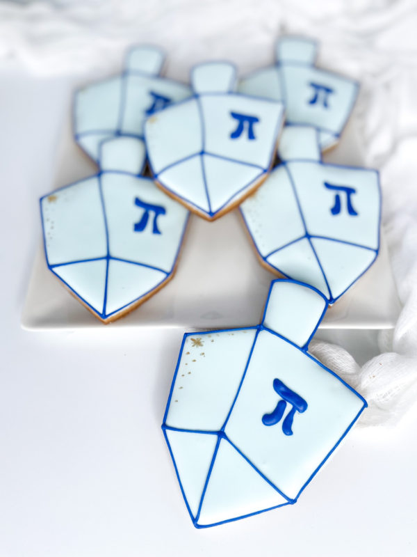 Light blue Dreidels write alt text Dreidel-shaped sugar cookies decorated with light blue and white icing, dark blue outlines, and the Hebrew letter He in the center.