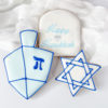 Hanukkah Gift Set **Alt Text:** Three decorated Hanukkah sugar cookies with white and light blue icing, including a dreidel, a Star of David, and an arch-shaped cookie reading "Happy Hanukkah."