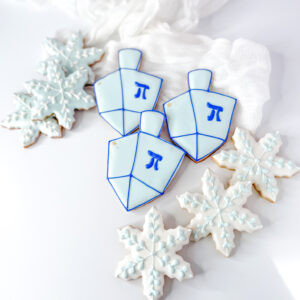 A top-down view of three dreidel-shaped sugar cookies decorated with light blue and royal blue icing alongside four snowflake-shaped sugar cookies with white and light blue detail, arranged on a plain white surface.