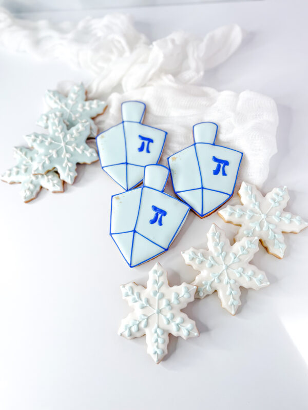 A top-down view of three dreidel-shaped sugar cookies decorated with light blue and royal blue icing alongside four snowflake-shaped sugar cookies with white and light blue detail, arranged on a plain white surface.