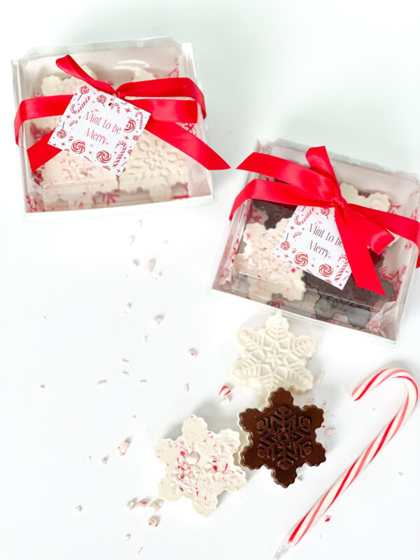 [Snowflake-shaped chocolates and peppermint barks packaged in clear gift boxes tied with red ribbons, displayed alongside loose snowflake treats, candy canes, and peppermint sprinkles]