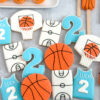 personalized basketball 2nd birthday cookies Basketball themed decorated sugar cookies including orange basketball cookies, basketball hoop cookies, court cookies, and personalized blue jersey cookies with name and number 2 for a basketball birthday party.