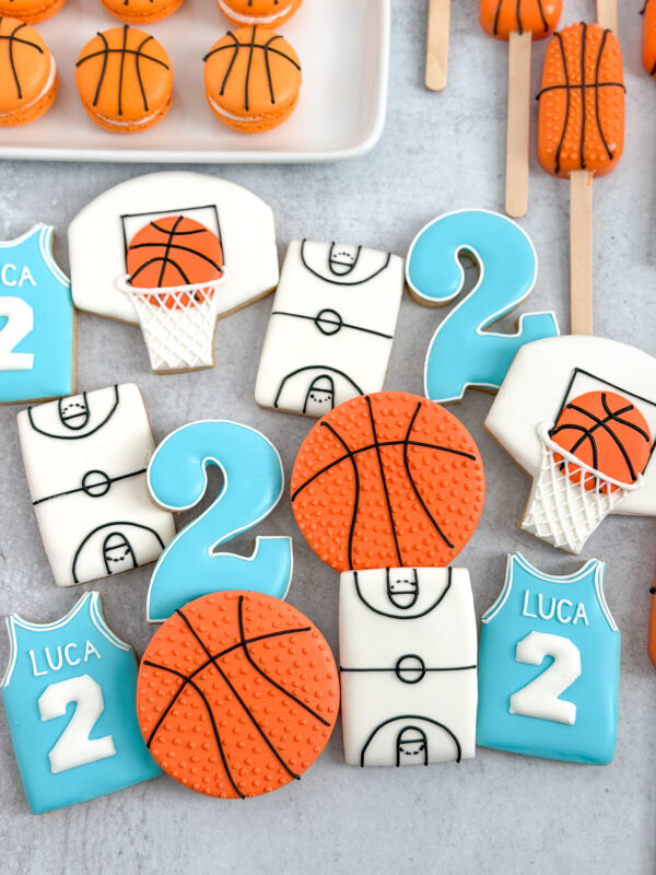 personalized basketball 2nd birthday cookies Basketball themed decorated sugar cookies including orange basketball cookies, basketball hoop cookies, court cookies, and personalized blue jersey cookies with name and number 2 for a basketball birthday party.