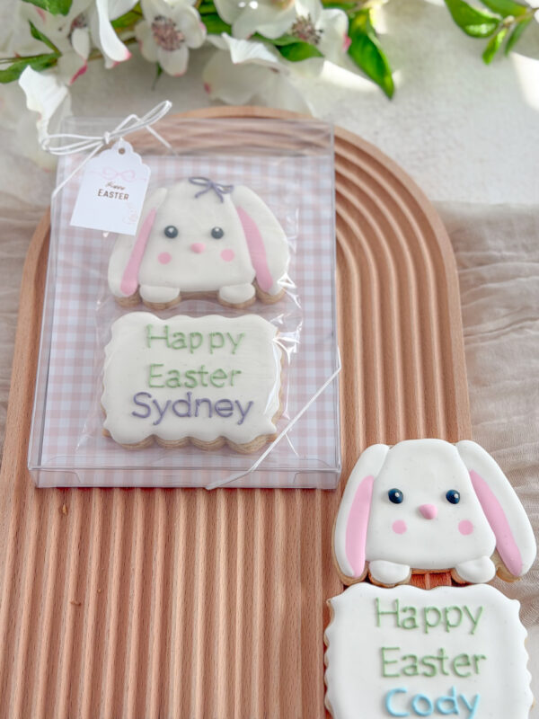 Hand-decorated Easter bunny sugar cookie set with a personalised name plaque, packaged in a clear gift box on a wooden tray with soft pastel decorations.