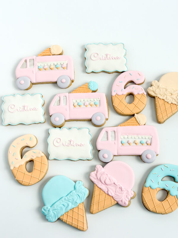 Hand-decorated pastel sugar cookies shaped like ice cream trucks, waffle cones, and number six cookies, with personalized name plaques , arranged on a light background.