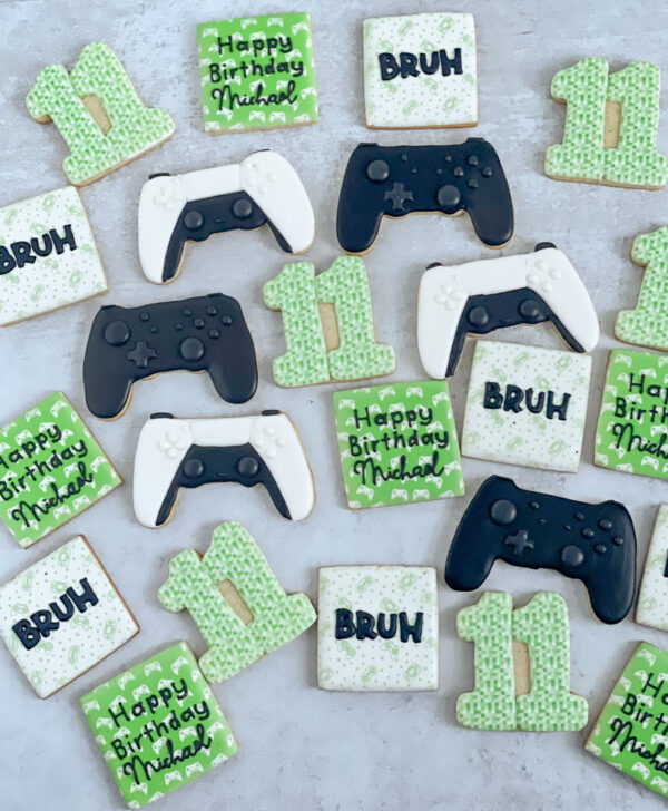 Video Game-theme birthday cookies black gaming controller, white and black gaming controller, number cookie with green pixelated design, square bruh cookie with gaming controllers background and happy birthday cookie and name