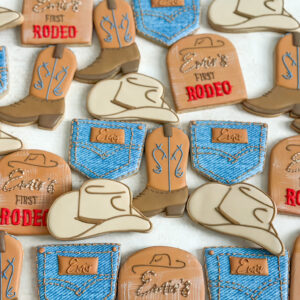 Custom Western rodeo sugar cookies decorated with cowboy boots, cowboy hats, denim jeans, and personalized rodeo details in blue, cream, caramel, and red.