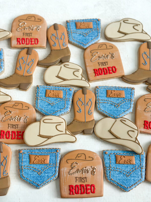 Custom first rodeo sugar cookies featuring cowboy boots, hats, denim jeans, and personalized details. Custom Western rodeo sugar cookies decorated with cowboy boots, cowboy hats, denim jeans, and personalized rodeo details in blue, cream, caramel, and red.