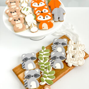 Decorated woodland baby shower cookies featuring adorable fox, bear, and raccoon cookies, leafy branch designs, baby onesies, and personalized “baby” script cookies in warm orange, brown, green, and soft gray icing.
