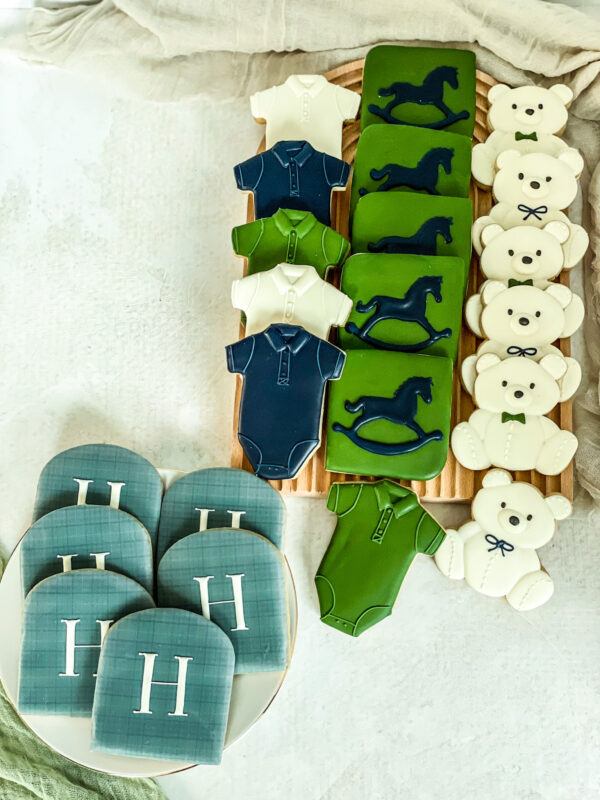 Custom baby boy sugar cookie set featuring teddy bears, rocking horses, baby bodysuits and personalized H monogram cookies in navy blue, green, & white.