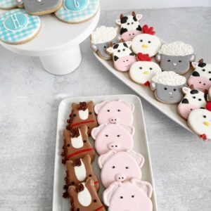 Close-up of hand-decorated farm animal sugar cookies with pigs, cows, sheep, chickens, and personalized blue gingham cookies arranged on white serving platters