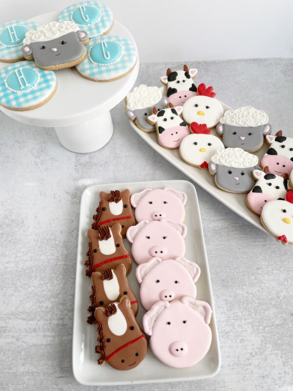 Custom farm animal sugar cookie set featuring pink pig, cow, sheep and chicken cookies with blue gingham monogram cookies Close-up of hand-decorated farm animal sugar cookies with pigs, cows, sheep, chickens, and personalized blue gingham cookies arranged on white serving platters